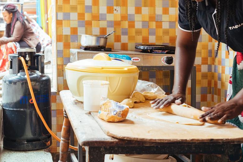 A person kneading dough on a wooden table in a kitchen with a yellow rice cooker, a gas stove, a large blue propane tank, and baking ingredients on the counter.