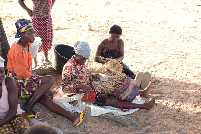 Women sit on the sandy ground weaving a straw basket, using colorful threads; one wears a patterned headwrap, another an orange blouse, and a younger person watches.