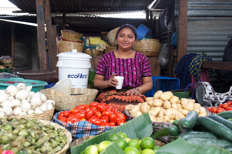 A smiling vendor sits behind a colorful display of fresh vegetables including tomatoes, potatoes, onions, cucumbers, and green beans at a market stall.