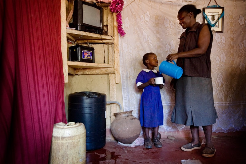 A woman pours water from a bright blue jug into a small white cup for a young girl in a blue dress, inside a modest, cluttered room with textured wallpaper.