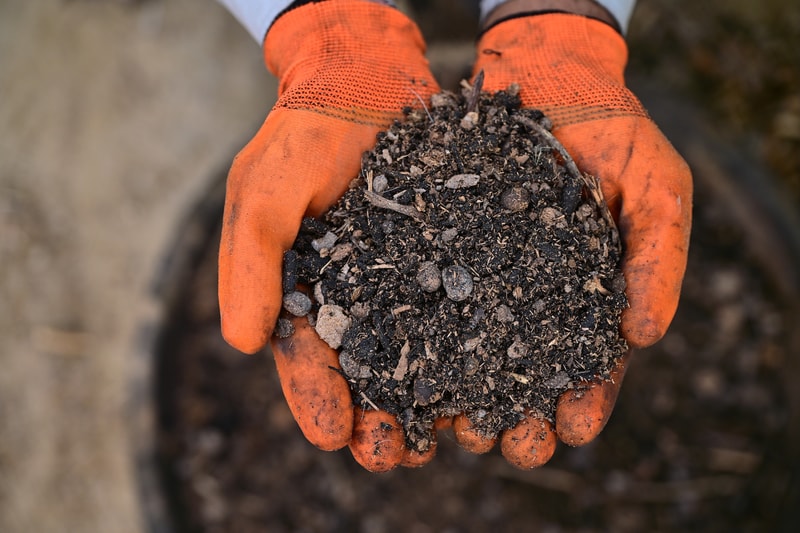 Two orange work gloves hold a handful of dark soil and small rocks, with gritty, clumpy earth and pebbles visible against a blurred background.