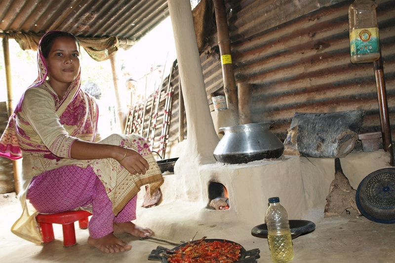 A woman in a traditional pink and cream sari sits on a small stool by a clay stove, cooking in a rustic corrugated‑metal shelter with chili peppers spread on a tray.