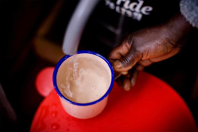 A dark-skinned hand grips a small white mug with a blue rim as hot water pours in, creating bubbles, on a red surface in a dim setting.