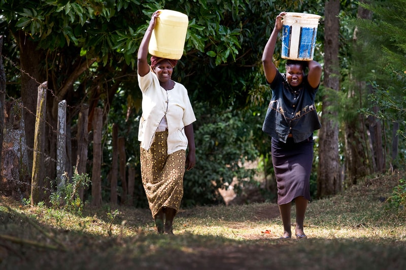 Two women walk along a dirt path carrying large buckets on their heads, one in a light blouse and patterned skirt, the other in a dark shirt and skirt, with greenery and trees in the background.