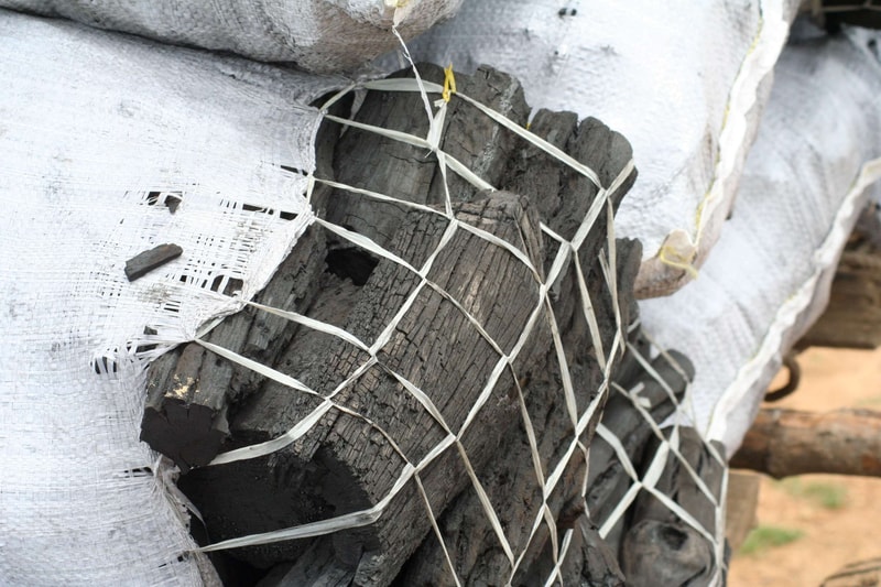 Bundled charred firewood wrapped in a white fabric cover and secured with a crisscross net, showing cracked blackened wood pieces peeking through.