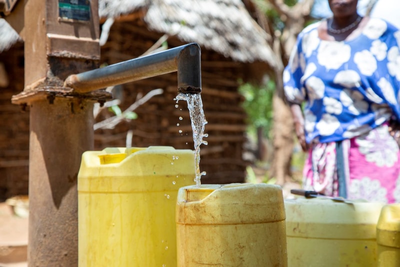 Water trickling from a metal spout into yellow plastic containers outdoors; a person in a blue floral garment stands nearby in a rural setting.