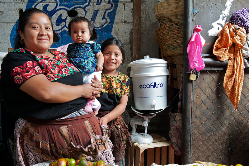 Three people stand in a market stall: a smiling woman wearing a floral blouse holds a baby, and a young girl in a traditional embroidered dress sits beside them; a white ecofiltro water filter sits on a stand to their right, with baskets, fabrics, and a brick wall in the background.
