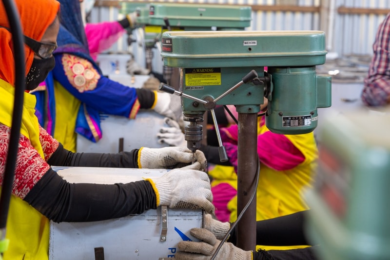 People wearing high-visibility clothing and gloves operate a green industrial drill press in a busy workshop.
