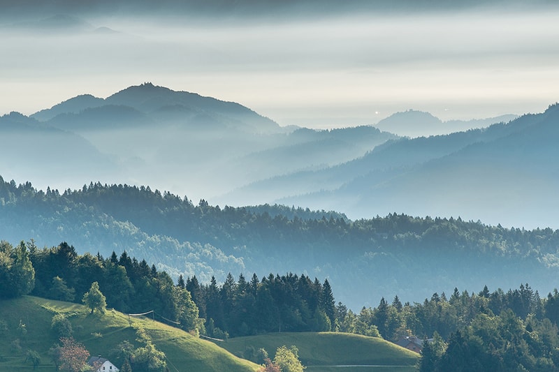 Layered misty blue mountains rise over rolling green hills and forests, with soft morning light and distant hazy valleys creating a tranquil scenic panorama.