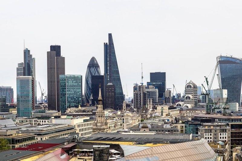 A panoramic city skyline featuring modern skyscrapers, including the Gherkin and The Shard-like tower, with dense mid-rise buildings and construction cranes.