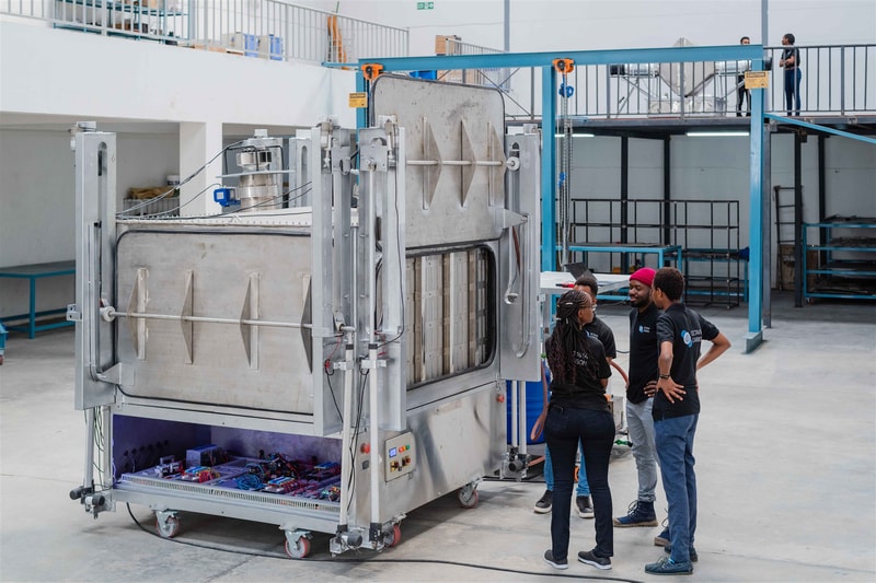 Three people wearing matching shirts stand in a workshop, discussing a large industrial stainless steel machine with a front access door and wheels, cables, and control panel.