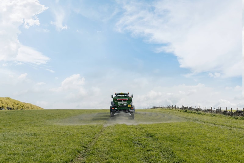 Large green agricultural tractor driving away on a grassy field, kicking up dust, with a wooden fence line along a rural path and a bright blue sky with scattered clouds.