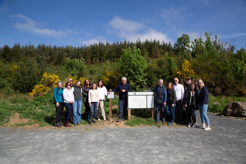 Group of people standing outdoors on a sunny day beside a signboard and map, with trees and shrubs in the background.