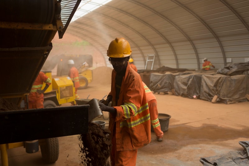 Workers in orange safety gear and hard hats operate a large industrial machine in a dusty, curved-roof facility, with piles and tarps in the background.