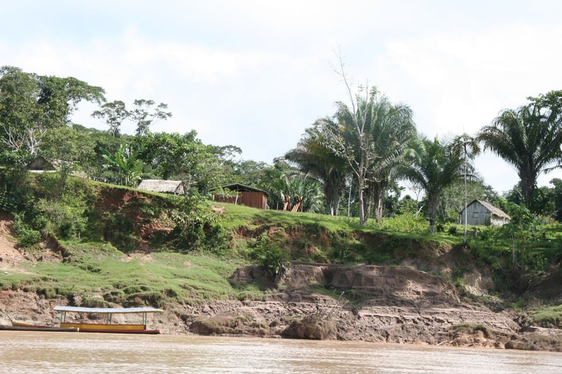 Hilly riverside scene with green vegetation, palm trees, small rustic huts, and a wooden boat on the muddy water by eroded clay banks under a partly cloudy sky.