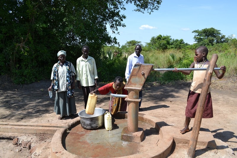 A group of people gathers around a hand-pumped water well in a rural area; one person pours water from a yellow container into a large metal pot while others watch.