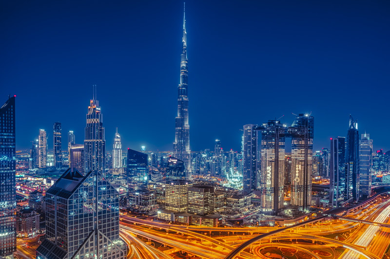 A nighttime cityscape with towering skyscrapers, including a tall illuminated central spire, bright building lights, and intertwined highway ribbons of golden traffic below.
