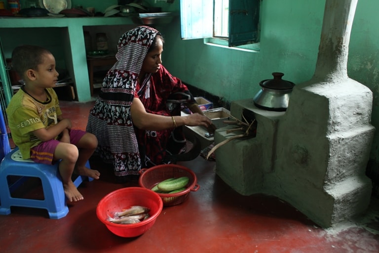 A woman wearing a patterned headscarf sits by a stove, preparing fish with a young child seated nearby on a blue plastic stool, two bowls of fish and vegetables nearby.