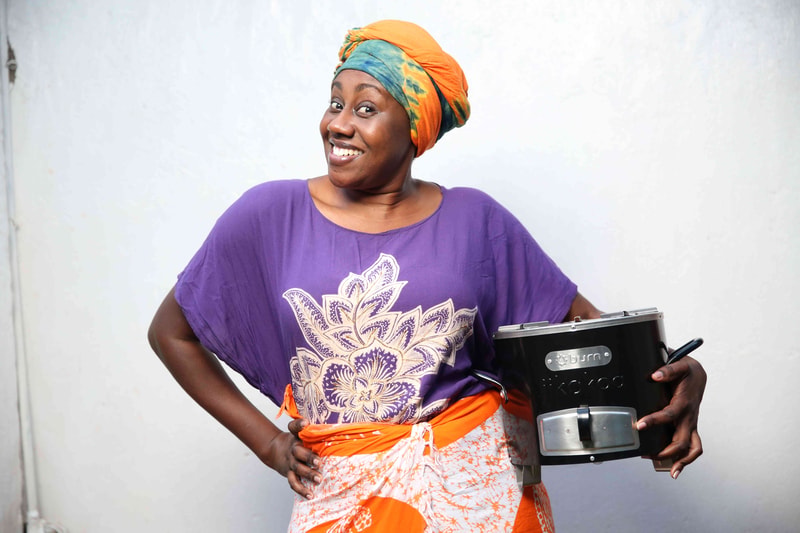 A smiling woman wearing a purple top and colorful head wrap holds a black kitchen appliance (blender) against a light textured wall, with an orange patterned wrap around her waist.
