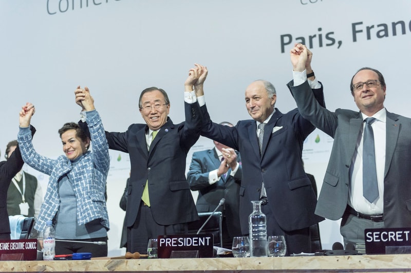 Group of diverse officials on stage holding hands up in unity, smiling, seated at a conference with nameplates like PRESIDENT and SECRETARY in front.