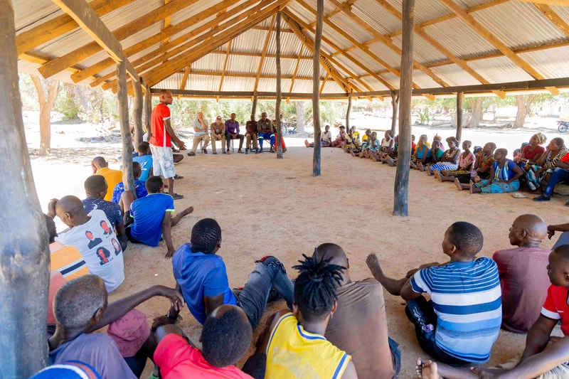 A large group of people sit in a circular arrangement inside an open, thatched pavilion, listening to a person in a red shirt speaking at the center.
