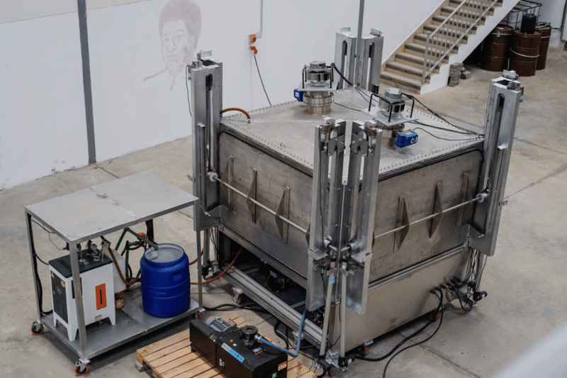 Industrial stainless steel apparatus with a large rectangular vessel, hydraulic clamps, electrical control box, blue drum, hoses, and a stairwell in the background.