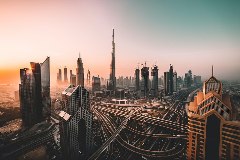 Sunlit cityscape with towering skyscrapers and a complex network of elevated highways, including the central Burj Khalifa silhouette amid a warm sunset glow.