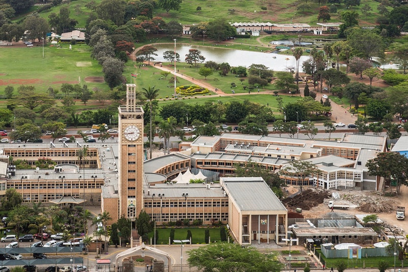 Aerial view of a large campus with a tall clock tower, surrounding buildings, a central green park, trees, a lake, and roads with parked cars.