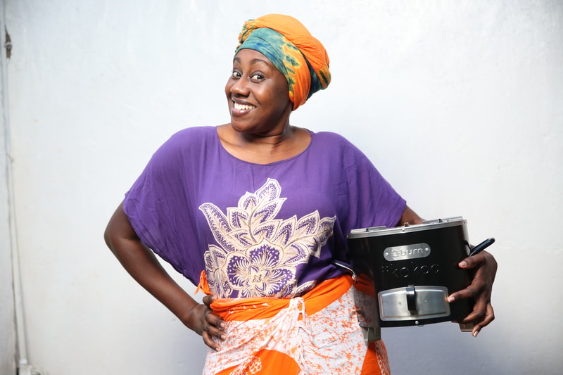 Smiling person wearing a purple top with a large white floral design, an orange and teal head wrap, and an orange patterned wrap, holding a black kitchen appliance.