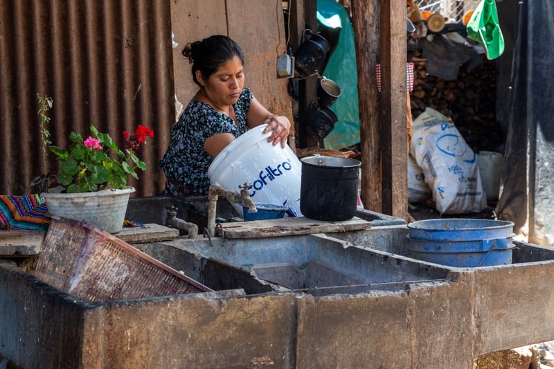 A woman in a patterned dark top rinses a white plastic bucket at an outdoor sink, with metal taps, a black pot, blue buckets, and rustic surroundings including wood and corrugated metal.