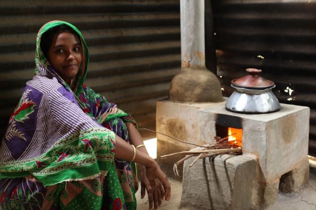 A woman wearing a colorful green and purple patterned sari sits beside a small brick stove with a fire and a metal pot, inside a simple shelter.