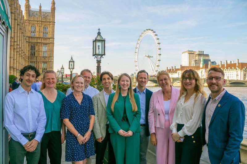 Group of ten adults posing outdoors by the Thames with the London Eye and Big Ben skyline, smiling in colorful smart casual attire on a sunny day.
