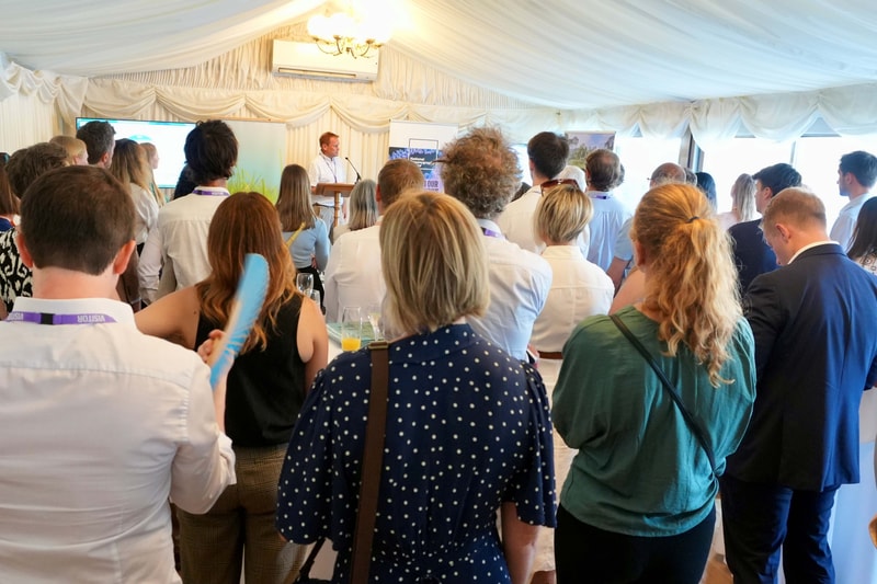 Crowd of people facing a speaker at a podium in a bright, white tent; banners and screens behind, attendees wearing lanyards and business casual attire.