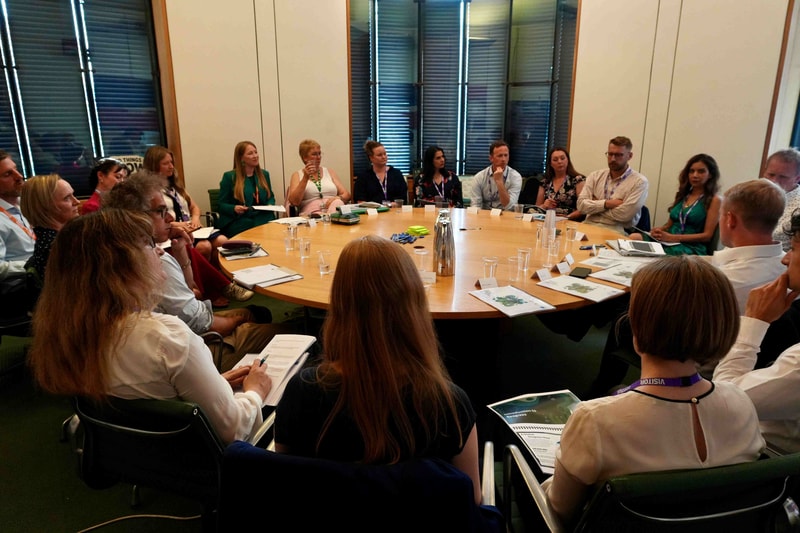 A diverse group of professionals sit around a large circular table in a conference room, discussing with papers, notebooks, and name placards scattered across the table.