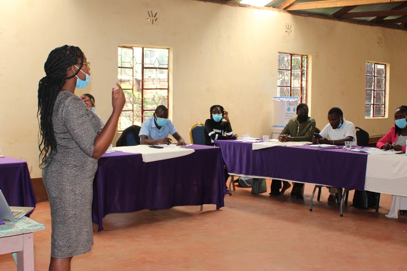 A woman wearing a mask stands at the left, speaking to a seated group around a long table covered with purple and white cloths in a sunlit room with windows.