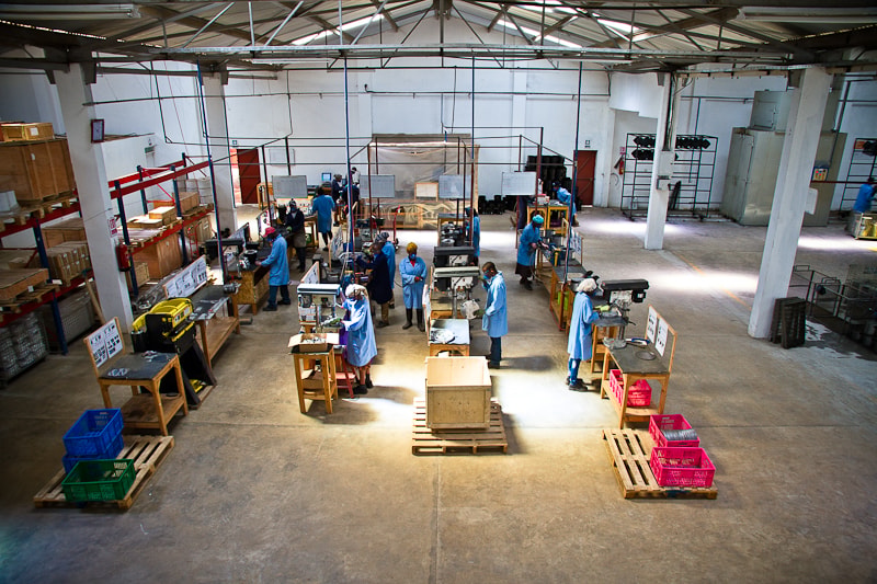 Workers in blue protective gowns and hairnets operate machinery at stations in a large industrial workshop, with organized workbenches, pallets, and storage shelves throughout.