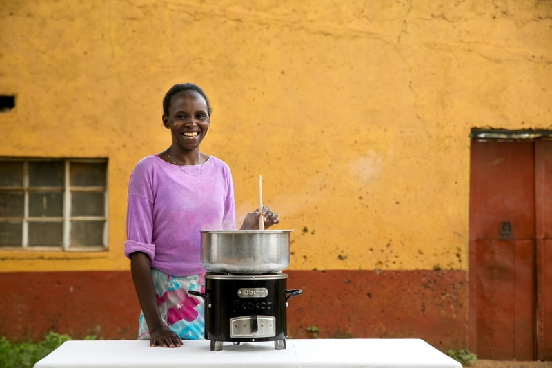 Smiling person wearing a purple long-sleeve top and patterned skirt stands beside a large stainless pot on a small black cooker, set against a yellow wall.