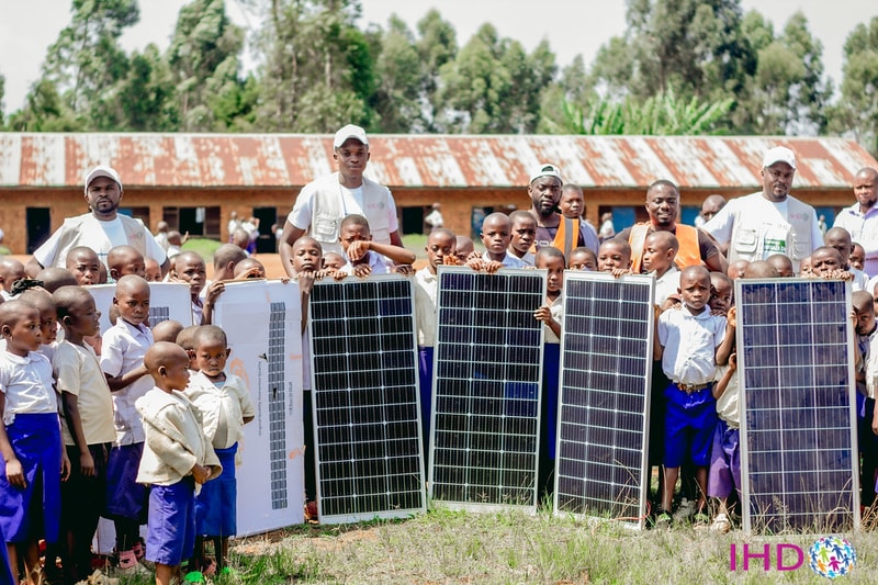 Group of children and volunteers posing outdoors with several large solar panels, in front of a rustic building and green trees.