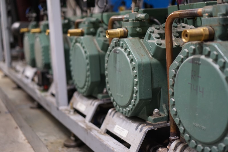 Industrial machinery with large green gear housings and copper piping, arranged in a row along a metal frame, slightly blurred perspective.