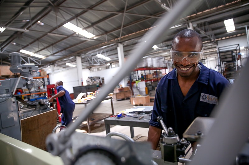 A smiling worker wearing safety goggles and a navy jumpsuit operates a metal machine in a spacious industrial workshop with other equipment and workers in the background.