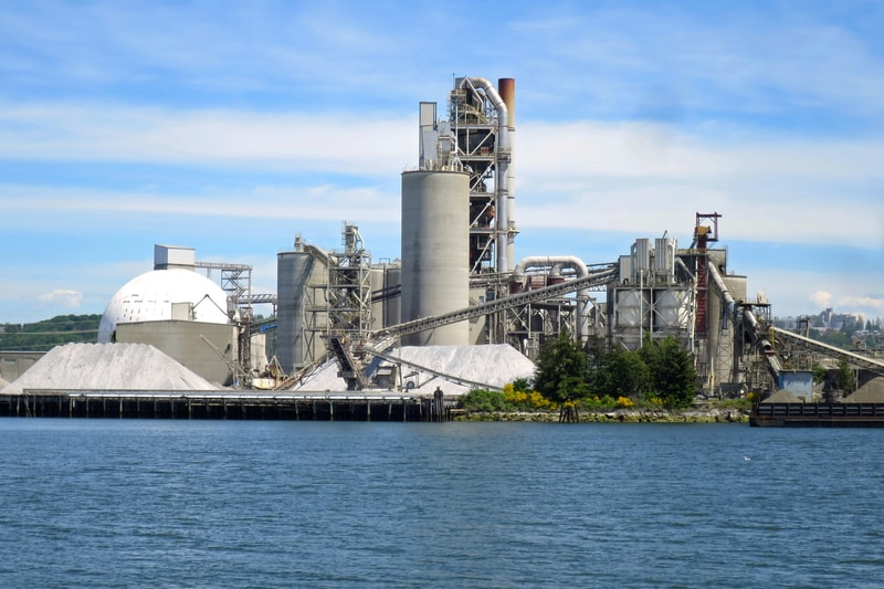 Industrial cement plant by the water, with tall silos, conveyors, and a large white dome, set against a blue sky and calm harbor foreground.