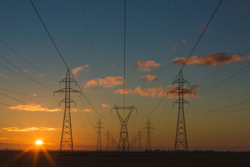 Power lines and tall steel lattice transmission towers stretch across a flat landscape at sunset, with orange glow on the horizon and scattered clouds.