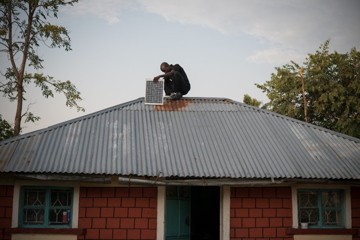 A person sits on a corrugated metal roof beside a solar panel, perched near the ridge of a single-story brick house with green-trimmed windows and a door, trees in the background.