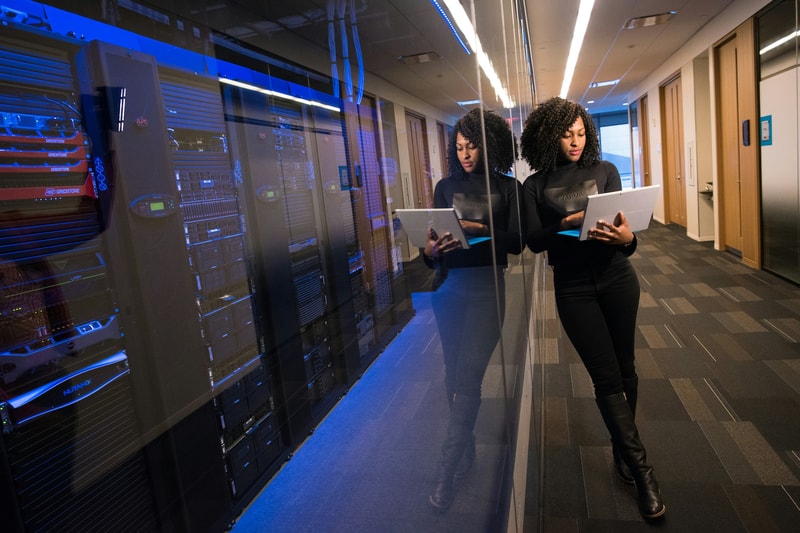 Two women with curly hair in black outfits stand in a server room museuming tablets, reflected in a glass wall, blue-lit racks and rows of servers beside.