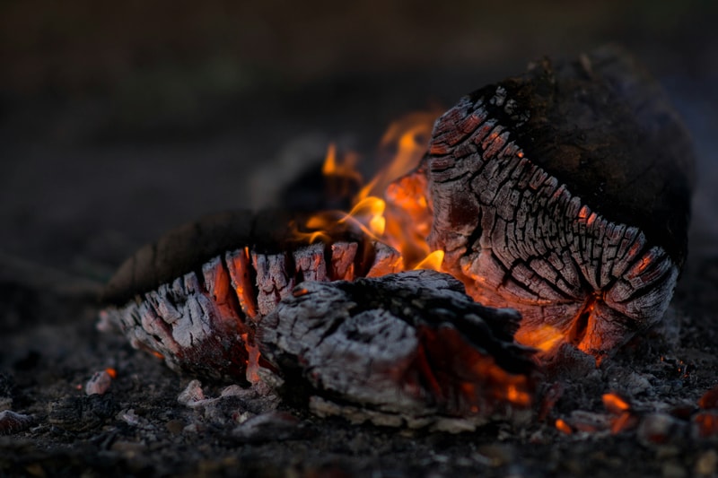Glowing embers and charred logs in a small campfire, with orange flames licking cracked, burnt wood and dark, smoky surroundings.