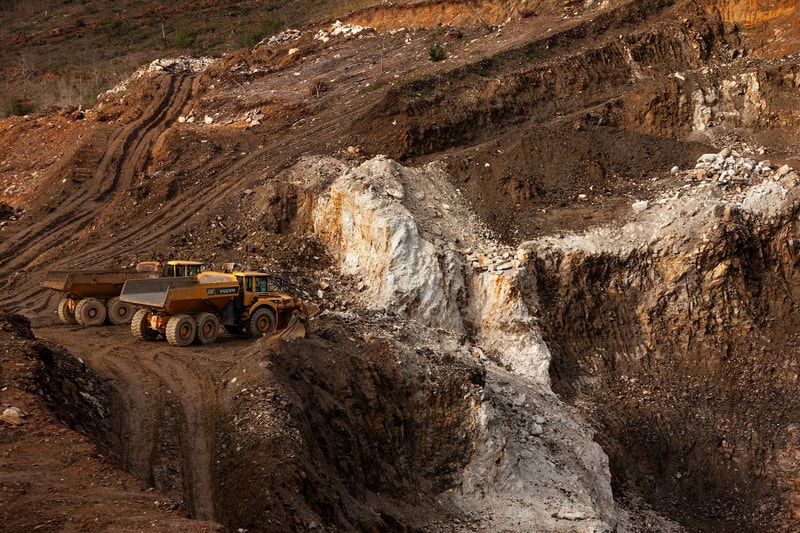 Open-pit mining site with large earth excavations and terraced brown soil slopes; two yellow Volvo dump trucks parked on dirt road among rocky terrain.