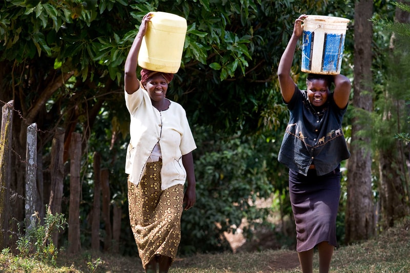 Two women walk along a dirt path in a leafy area, each balancing a large container on their heads—one yellow, the other a weathered blue-and-white bucket.