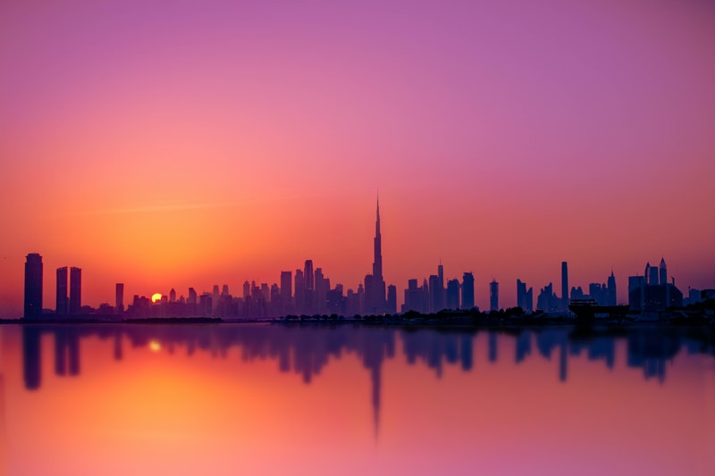 Silhouette of a modern city skyline at sunset with tall central tower, reflected in calm water, sky fading from orange to pink and purple.
