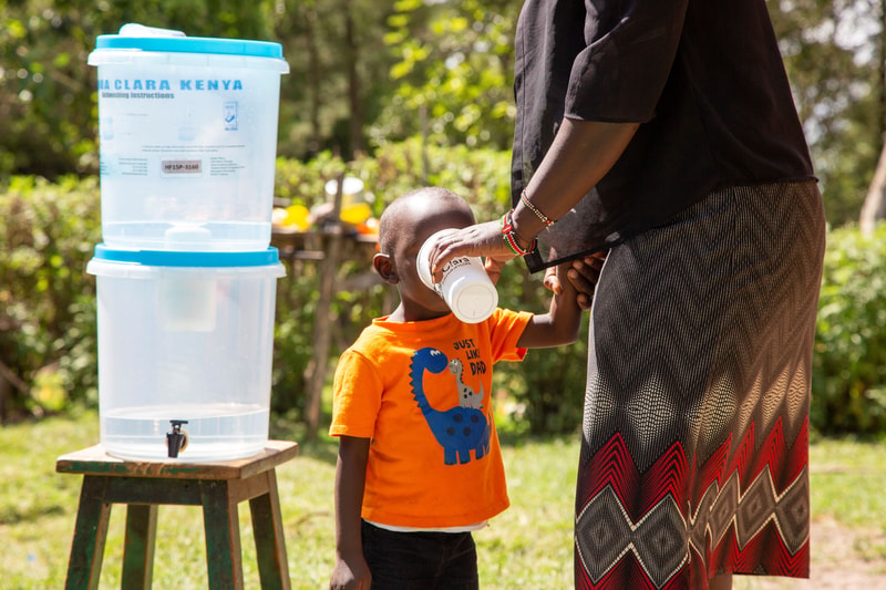 A child in an orange shirt drinks from a white cup held by an adult outdoors, beside a stacked water dispenser on a wooden stool in a sunny setting.