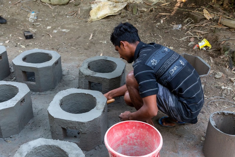 A man in a striped shirt crouches and smooths cement around cylindrical concrete blocks outdoors, with a red bucket nearby and construction debris scattered on the ground.